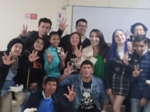 Grupo de jóvenes reunidos en un salón, sonriendo y haciendo el signo de la paz con las manos.